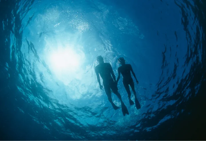 Two snorkelers silhouetted underwater beneath bright sunlight in deep blue ocean water