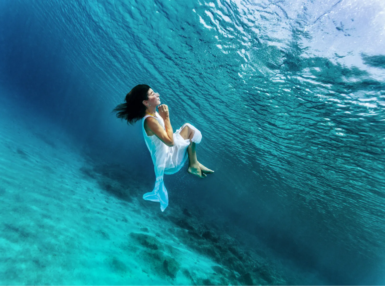 A person in a white dress floating underwater above a sandy ocean floor
