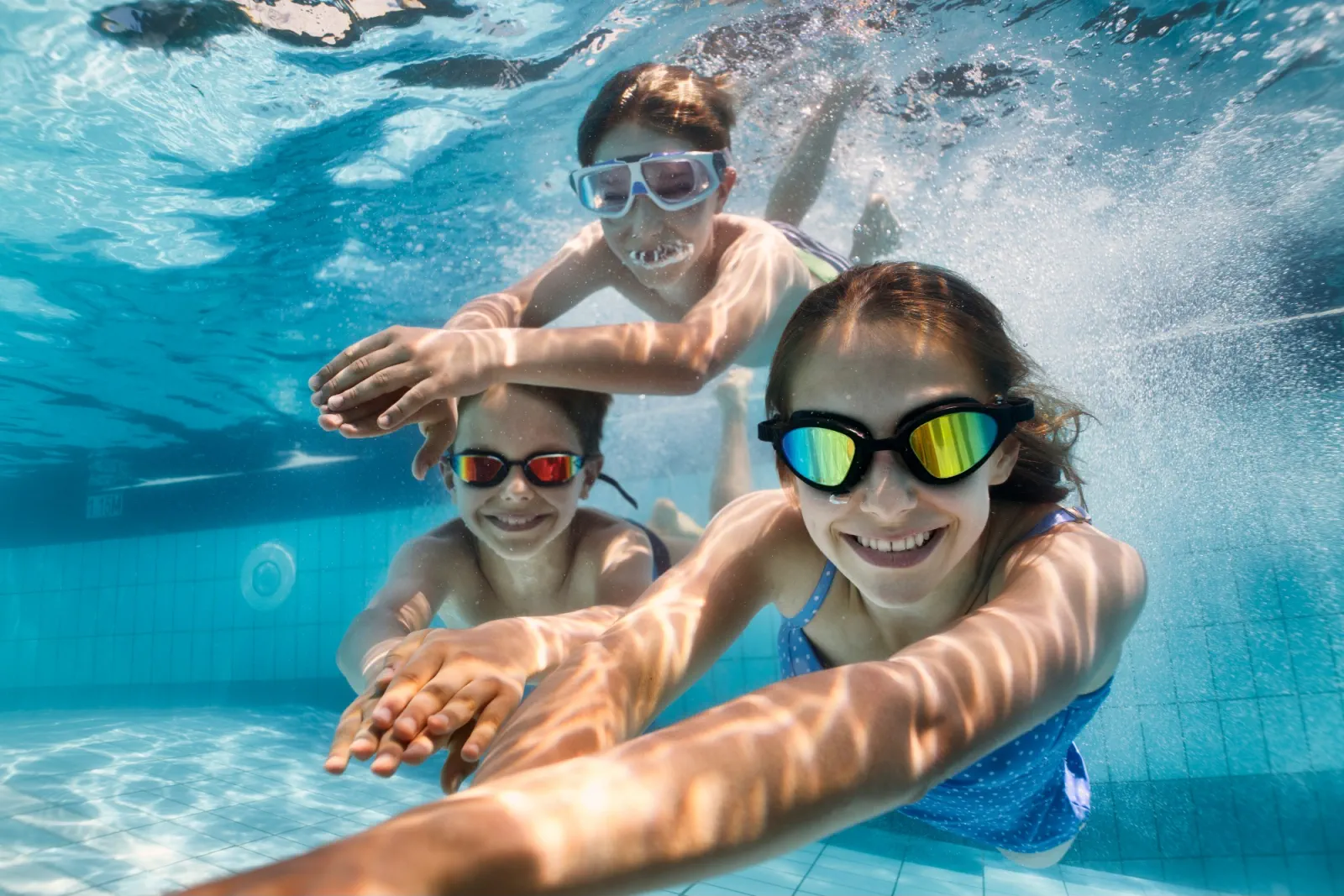 Three young swimmers smiling underwater in a bright pool with goggles